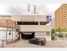 Außenansicht eines mehrstöckigen Parkhauses mit der Beschriftung „Q-Park Märkische Zeile“. Ein blauer Hinweis mit einem „P“-Symbol signalisiert Parkmöglichkeiten, und der Eingang ist durch eine Schranke markiert. Im Vordergrund fahren Autos, während rechts ein gelbes Wohngebäude und grüne Bäume sichtbar sind.