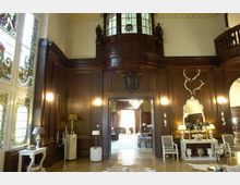 Großer Raum im historischen Stil mit dunklen Holzvertäfelungen, verzierten Buntglasfenstern und einer dekorativen Holzbalkon-Nische. Der Raum ist mit antiken Möbeln, Spiegeln, Lampen und Jagdtrophäen dekoriert, und im Hintergrund ist ein angrenzender Raum sichtbar.