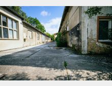 Eine verlassen wirkende Hofanlage mit alten, teilweise verfallenen Gebäuden aus Putz und Ziegeln. Der gepflasterte Hof ist von Bäumen und Sträuchern umgeben, die teils an den Wänden hochwachsen.