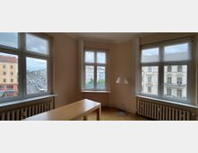 Heller, leerer Wohnraum mit hellen Wänden, Holzfußboden und großformatigen Eckfenstern auf zwei Seiten sowie einem weiteren Fenster an der Stirnseite. Vor den Fenstern stehen Heizkörper; im Raum befinden sich ein schlichter Holztisch und eine Stehlampe, mit Blick auf eine städtische Straße und gegenüberliegende Altbaufassaden.