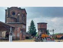 Historisches Bahngelände mit Backsteinbauten, darunter ein hohes, quadratisches Gebäude mit runden Fenstern und ein achteckiger Wasserturm. Im Vordergrund sind Gleisanlagen mit einem alten gelben Zug sowie weiteren technischen Anlagen zu sehen, umgeben von einer teils bewachsenen Umgebung.