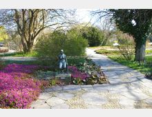 Ein Gartenweg aus Steinplatten führt durch eine parkähnliche Landschaft mit Büschen, großen Bäumen und violett blühenden Sträuchern. Im Vordergrund steht eine bronzene Skulptur einer knienden Figur auf einem Sockel.