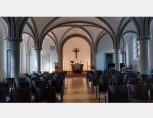 Innenraum einer kleinen Kirche mit gotischen Bögen und grauen Säulen. Im Vordergrund sind Stuhlreihen, im Hintergrund ein schlichter Altar mit Kreuz, Kerzen und Blumenschmuck auf einem erhöhten Podest.