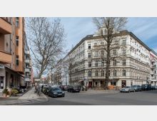 Straßenecke in einem städtischen Wohngebiet mit altmodischen Mehrfamilienhäusern im Gründerzeitstil; links orangefarbene Gebäudefassade mit Balkonen, rechts ein weißes, reich verziertes Eckgebäude. Parkende Autos und kahle Bäume säumen die Straße.