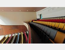 Ein moderner Vorlesungssaal mit ansteigender Bestuhlung in verschiedenen Farben (gelb, rot, und schwarz). Die Wände bestehen aus glatten, cremefarbenen und hölzernen Elementen, und die Decke ist mit eingebauten Leuchten ausgestattet.