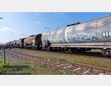 Ein Güterzug mit mehreren braunen und grauen Kesselwaggons, teilweise mit Graffiti, steht auf Gleisen in einem Bahnhofsbereich. Im Hintergrund sind weitere Gleise, Lampenmasten und offener Himmel zu sehen.