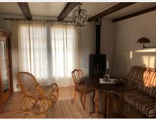 Wohnzimmer mit rustikalem Stil; ein Holzschaukelstuhl, ein runder Holztisch mit Stühlen und eine Couch mit gestreiftem Bezug stehen auf einem Holzboden. Im Hintergrund befinden sich bodenlange Vorhänge vor großen Fenstern, ein Kaminofen und eine hängende Kristalllampe.