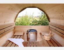 Innenansicht einer holzvertäfelten Fasssauna mit leicht gewölbter Decke, Sitzbänken aus Holz auf beiden Seiten und einem Saunaofen aus Edelstahl in der Mitte. Durch ein großes halbrundes Fenster ist eine grüne Landschaft mit Bäumen sichtbar.