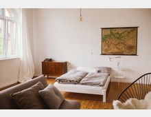 Helles Schlafzimmer mit einem schlichten weißen Bett, grauer Bettwäsche und einem Nachttisch mit Lampe. An der weißen Wand hängt eine gerahmte Karte, daneben steht ein Holzschrank mit Schreibmaschine; das Zimmer hat Holzboden und ein großes Fenster mit weißen Vorhängen.