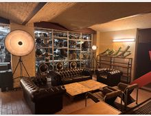 Industrie-inspirierter Raum mit freiliegenden Ziegelwänden, braunen Ledersofas im Chesterfield-Stil und einem niedrigen Holztisch. Im Hintergrund stehen Regale mit vintage Bühnen- und Filmbeleuchtung sowie ein leuchtendes Schriftzeichen "seit".