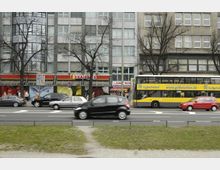 Straßenszene mit mehreren Autos und einem gelb-blauen Linienbus auf einer mehrspurigen Straße vor einer Häuserfassade mit Geschäften, darunter ein Supermarkt mit dem Logo "Kaiser's". Die Gebäude sind von kahlen Bäumen umgeben, und im Vordergrund befindet sich ein schmaler Grünstreifen.
