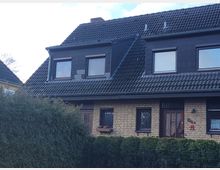 Zweistöckiges Wohnhaus mit Satteldach und Dachgauben, verkleidet mit dunklen Dachziegeln. Die Fassade im Erdgeschoss ist aus gelbem Klinker, mit mehreren Fenstern und einer Holztür; davor eine grüne Hecke.