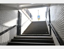 Treppenaufgang in einer U-Bahn-Station, umgeben von Wänden aus hellen Kacheln. Am oberen Ende der Treppe ist ein Schild mit einem blauen „U“ zu sehen, das auf die U-Bahn hinweist.
