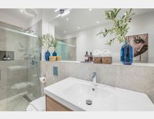 Modernes Badezimmer mit großer, begehbarer Glasdusche, hellgrauen Wandfliesen und einem breiten Waschtisch mit Spiegelwand und Einbaubeleuchtung. Auf der Ablage stehen blaue Glasvasen mit Zweigen, Körbe mit Handtüchern sowie Seifenspender; an der Wand hängt ein dekoratives Bild.