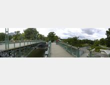 Eine Fußgängerbrücke mit grün gestrichenen Geländern und dekorativen Laternen erstreckt sich über einen Fluss, umgeben von Bäumen und Grünflächen. Im Hintergrund sind ein moderner Gebäudekomplex und weitere Parkwege zu sehen.