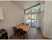 Heller Essbereich in einem modernen Holzhaus mit weiß vertäfelten Wänden, Holzboden und Esstisch mit vier Stühlen. Große bodentiefe Glas-Schiebetür mit Oberlicht und weißen Vorhängen führt auf eine Holzterrasse mit Blick ins Grüne.