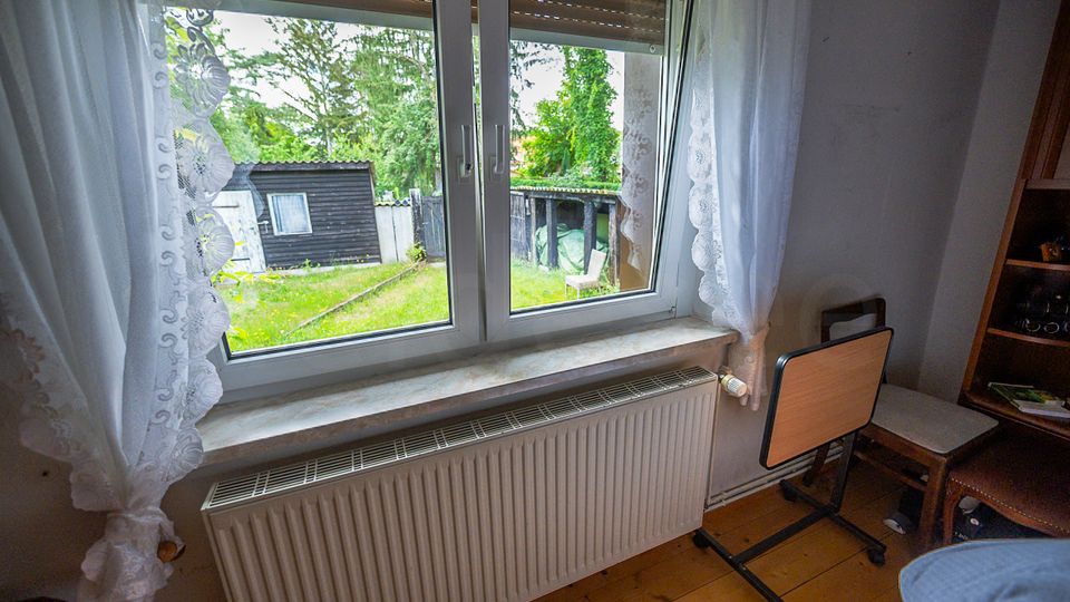 Blick aus einem kleinen Zimmer durch ein zweiflügeliges Fenster mit Spitzenvorhängen und Rollladen auf einen Garten mit Rasen, Schuppen und Zaun. Unter dem Fenster befindet sich ein Heizkörper; im Raum sind ein Stuhl und ein Regal zu sehen.