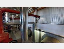 Innenansicht einer Industrieanlage mit großen Rohren in Metall- und Rotlackoptik. Die Umgebung zeigt eine Halle mit gewelltem Blechdach, weißen Wänden, Leitern und technischen Geräten.