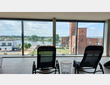 Innenansicht eines Raumes mit großen bodentiefen Fenstern, die den Blick auf eine Flusslandschaft mit Bäumen, einer Brücke und einem roten Backsteingebäude freigeben. Im Vordergrund stehen moderne Liegestühle und kleine Beistelltische auf einem gefliesten Boden.