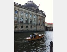 Historisches Gebäude mit Kuppeldach am Ufer eines Flusses, im Vordergrund ein hölzernes Motorboot mit mehreren Personen, das auf dem Wasser fährt.