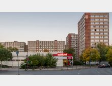 Straßenszene mit Wohnhochhäusern im Hintergrund, die eine typische Plattenbau-Architektur aufweisen. Im Vordergrund befindet sich ein kleiner Supermarkt mit dem Namen "Nahkauf", umgeben von Bäumen und vereinzelten parkenden Autos.
