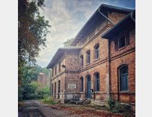 Ein historisches Backsteingebäude mit Rundbogenfenstern und einem Schild mit der Aufschrift „Fahrkarten-Ausgabe“. Der Vordergrund zeigt einen gepflasterten Weg, umgeben von Bäumen und Grünflächen, während im Hintergrund ein weiteres Gebäude sichtbar ist.