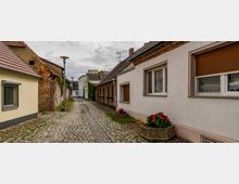 Schmale gepflasterte Gasse mit niedrigen Häusern auf beiden Seiten, teils mit verputzten Fassaden, teils mit sichtbaren Backsteinwänden. Am Straßenrand stehen Blumenkübel mit farbigen Blumen, und ein bewölkter Himmel ist im Hintergrund sichtbar.