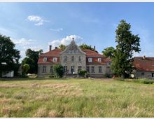 Historisches Gutshaus mit verputzter Fassade und roten Ziegeldächern, flankiert von zwei seitlichen Gebäudeflügeln. Das Gebäude steht inmitten einer Wiesenlandschaft mit einzelnen Bäumen, unter einem klaren blauen Himmel.