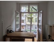 Ein schlichtes Zimmer mit weiß verputzten Wänden und einem Holztisch im Vordergrund. Im Hintergrund befinden sich geöffnete Doppelfenster, die auf einen kleinen Balkon führen, mit Blick auf Bäume und eine Wohnhausfassade gegenüber.