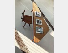 Ein minimalistischer Arbeitsplatz mit einem hölzernen Schreibtisch in asymmetrischer Form und einem passenden Holzstuhl auf hellem Betonboden. Auf dem Tisch liegen vier offene Bücher, während schräg einfallendes Sonnenlicht durch Fensterflächen Schattenstreifen erzeugt.