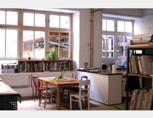 Ein heller Arbeitsraum mit großen Fenstern, einem Holztisch mit Stühlen in der Mitte und Bücherregalen entlang der Wand. Im Hintergrund befindet sich eine kleine Küchenecke mit einer Arbeitsplatte und Haushaltsgegenständen.