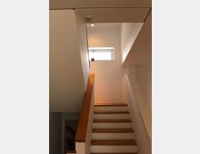 Treppenhaus mit modernem Design, weißen Wänden und Holzstufen. Oben befindet sich ein kleines rechteckiges Fenster, durch das Licht einfällt, mit einer kleinen Pflanze auf der Fensterbank.