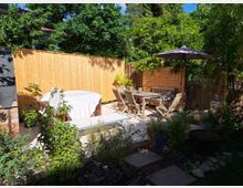 Ein gemütlicher Gartenbereich mit einer Holzterrasse, auf der ein Holztisch mit Stühlen und eine Bank unter einem Sonnenschirm stehen. Im Hintergrund befinden sich eine Sichtschutzwand aus Holz und grüne Bepflanzung, während im Vordergrund verschiedene Pflanzen und ein überdachter Whirlpool zu sehen sind.