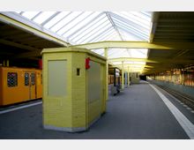 Ein überdachter U-Bahnhof mit gelben Backsteinwänden, einer gelben Stahlkonstruktion und einem gläsernen Dach. Im Hintergrund sind ein gelber Zug, Sitzbänke und Gleise sichtbar, die sich entlang eines gebogenen Bahnsteigs erstrecken.