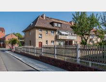 Straßenansicht eines Wohnhauses mit mehreren Wohnungen, hellbrauner Putzfassade und schrägem Ziegeldach. Vor dem Gebäude stehen Bäume, und ein Holzzaun sowie ein gemauerter Sockel säumen das Grundstück.