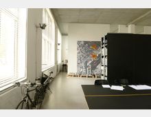Helles Büro mit großen Fenstern auf der linken Seite, die Tageslicht hereinlassen, einer Lagerregalwand rechts und einem großen Tisch mit Papieren im Vordergrund. An der Wand im Hintergrund hängt eine farbige Karte oder Grafik mit Stadtplan-Elementen, während links ein Fahrrad steht.