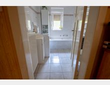 Blick durch eine Tür in ein hell gefliestes Badezimmer mit großer Badewanne an der Stirnwand und einem Fenster darüber. Links stehen weiße Badmöbel und ein hoher Schrank, rechts ist eine weitere Tür zu einem angrenzenden Bereich sichtbar.