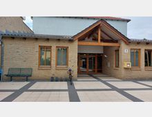 Eingangsbereich eines Backsteingebäudes mit Flachdach und Holzelementen, darunter ein holzüberdachtes Vordach. Vor dem Gebäude befinden sich ein Fahrradständer, eine Bank und eine gelbe Briefkastenanlage.