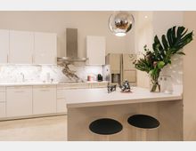 Moderne Küche mit weißen Hochglanzfronten, einer Marmor-Rückwand und einer Dunstabzugshaube aus Edelstahl. Im Vordergrund befindet sich eine Kücheninsel mit zwei schwarzen Hockern, einer Teekanne, Tassen und einem Glasvasen-Arrangement mit Blumen und grünen Blättern.