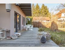 Holzterrasse eines modernen Hauses mit überdachtem Bereich, dekoriert mit Pflanzen in Töpfen, Laternen und einem Tisch mit Stühlen. Im Hintergrund ein Garten mit Rasen, kahlen Büschen und Bäumen sowie umliegenden Nachbarhäusern.