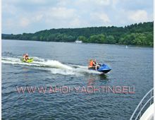 Zwei Personen fahren mit einem blauen Jetski, gefolgt von einem grünen Schlauchboot, über einen breiten See. Im Hintergrund sind ein bewaldetes Ufer und ein Boot sichtbar.