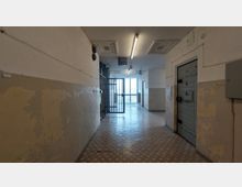 Ein heller Gefängnisflur mit abgeblätterter Wandfarbe, vergitterten Türen und vergitterten Fenstern am Ende des Ganges. Rechts befindet sich eine schwere Metalltür mit der Nummer "325".