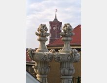 Zwei verzierte steinerne Balustersäulen mit kunstvollen Reliefs stehen im Vordergrund. Im Hintergrund ist das Rote Rathaus in Berlin mit seiner rot gefärbten Backsteinfassade und der Uhr auf dem Turm zu sehen; eine kleine Fahne weht an der Spitze des Turms.