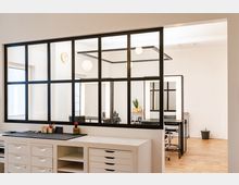 Ein heller Raum mit modernen Einrichtungselementen, darunter eine schwarze Glaswand mit Gittermuster, die einen Arbeitsbereich mit Spiegeln und Stühlen von einem vorderen Bereich mit weißen Schubladen trennt. Der Boden besteht aus hellem Holz, und an der Wand hängt eine runde Uhr.