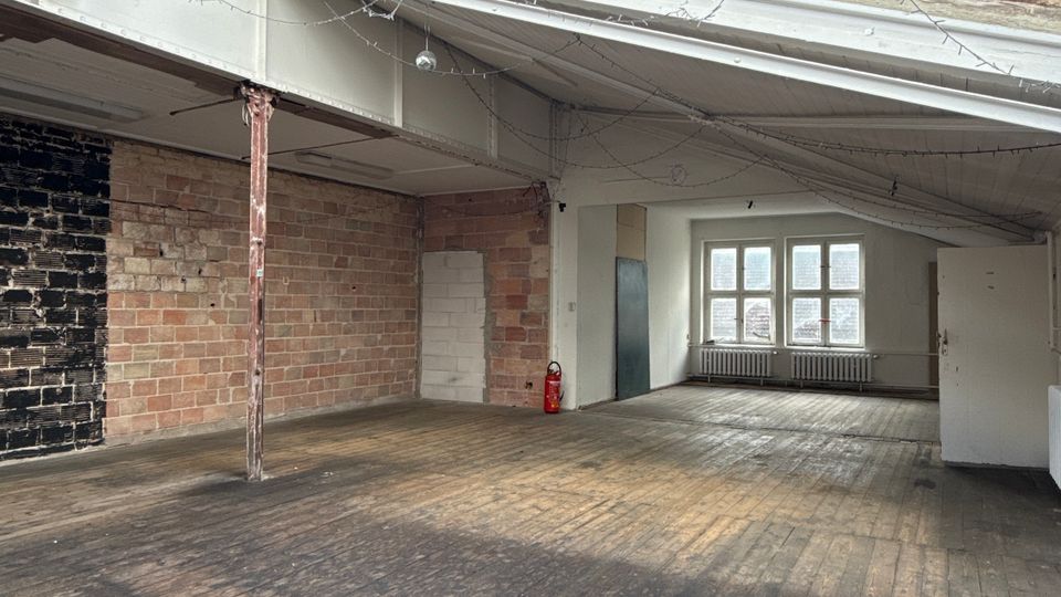 Großer, leerer Loft- oder Lagerraum mit abgenutztem Holzboden, Sichtziegelwänden und schräger Decke mit Oberlichtfenstern. An der Rückwand stehen mehrere rechteckige Fenster über einem Heizkörper, daneben eine Tür und ein Feuerlöscher.