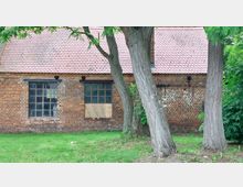 Eine Backsteinfassade eines alten Gebäudes mit zwei großen, vergitterten Fenstern, von denen eines teilweise mit Brettern vernagelt ist; darüber ein Dach mit roten Ziegeln. Im Vordergrund befinden sich mehrere große Bäume sowie eine Grünfläche.