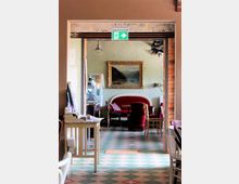 Innenraum eines gemütlichen Cafés mit gemustertem Boden in Grün- und Weißtönen. Im Hintergrund sind rote Polstermöbel, ein an der Wand hängendes Landschaftsgemälde in einem goldenen Rahmen und ein Notausgangsschild zu sehen.