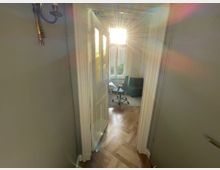 Ein Blick durch eine geöffnete weiße Tür in einen hellen Raum mit einem großen Fenster und einfallendem Sonnenlicht. Im Raum befinden sich ein Bürostuhl, ein Schreibtisch und eine Stehlampe, der Boden ist mit Parkett ausgelegt.