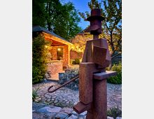 Metallskulptur in einem Garten mit Kopfsteinpflasterweg, umgeben von Pflanzen und beleuchteten Backsteingebäuden im Hintergrund während der Dämmerung.