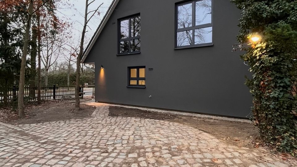 Ein modernes, dunkelgraues Einfamilienhaus mit Satteldach und großen Fenstern, umgeben von Bäumen und Hecken. Vor dem Haus befindet sich eine gepflasterte Einfahrt aus unregelmäßigen Steinplatten.